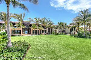 This image showcases the rear exterior of a luxurious property, featuring a well-manicured lawn that extends towards the building. Palm trees dot the landscape, adding a tropical feel, while the building itself has a modern architectural design with large windows and a warm color palette. The perspective is from the lawn looking towards the building, emphasizing the spaciousness and connection to the outdoors.