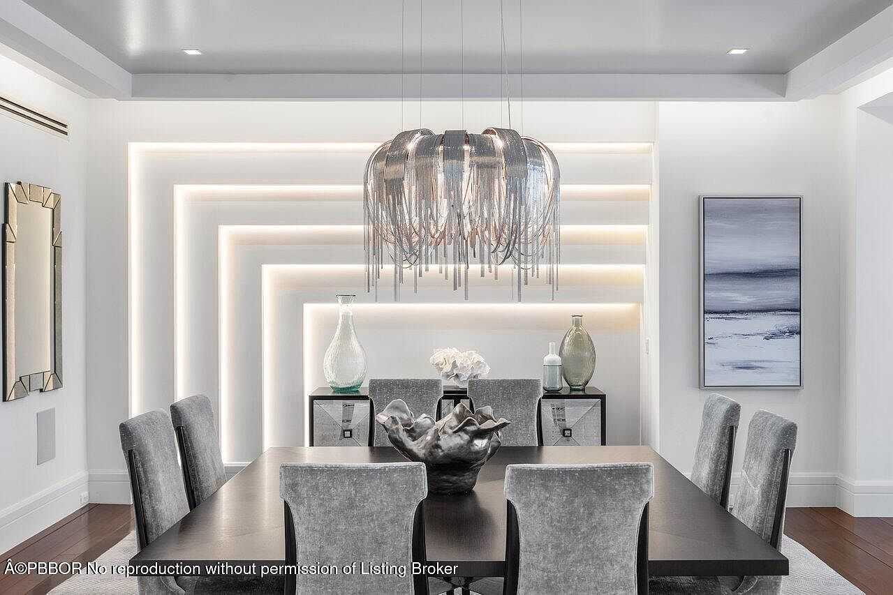 This is an interior shot of a modern dining room featuring a dark wood table surrounded by gray upholstered chairs. A striking chandelier hangs above the table, and the wall behind the table has a unique geometric design with integrated lighting. The room is decorated with vases and artwork, creating a sophisticated and elegant atmosphere.