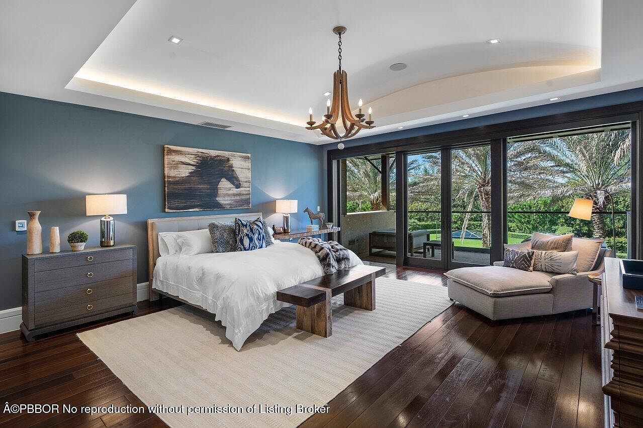 This is a primary bedroom featuring a king-size bed with white linens and decorative pillows, a wooden bench at the foot of the bed, and a dresser with lamps. The room has a large painting of a horse above the bed and sliding glass doors that lead to an outdoor area. The flooring is dark hardwood, and the walls are painted a calming blue, creating a serene and luxurious atmosphere.