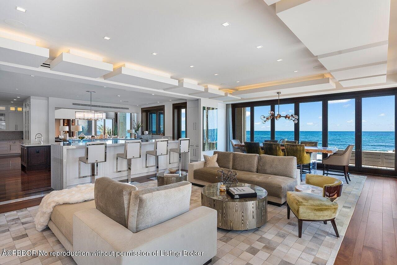 This is a luxurious open-concept living space featuring a modern kitchen with bar seating, a comfortable living area with plush sofas and a round coffee table, and a dining area with ocean views. The room is well-lit with recessed lighting and a statement chandelier, and the large windows offer stunning views of the ocean, creating a serene and upscale atmosphere. The perspective is from a low angle, emphasizing the spaciousness and high-end finishes.