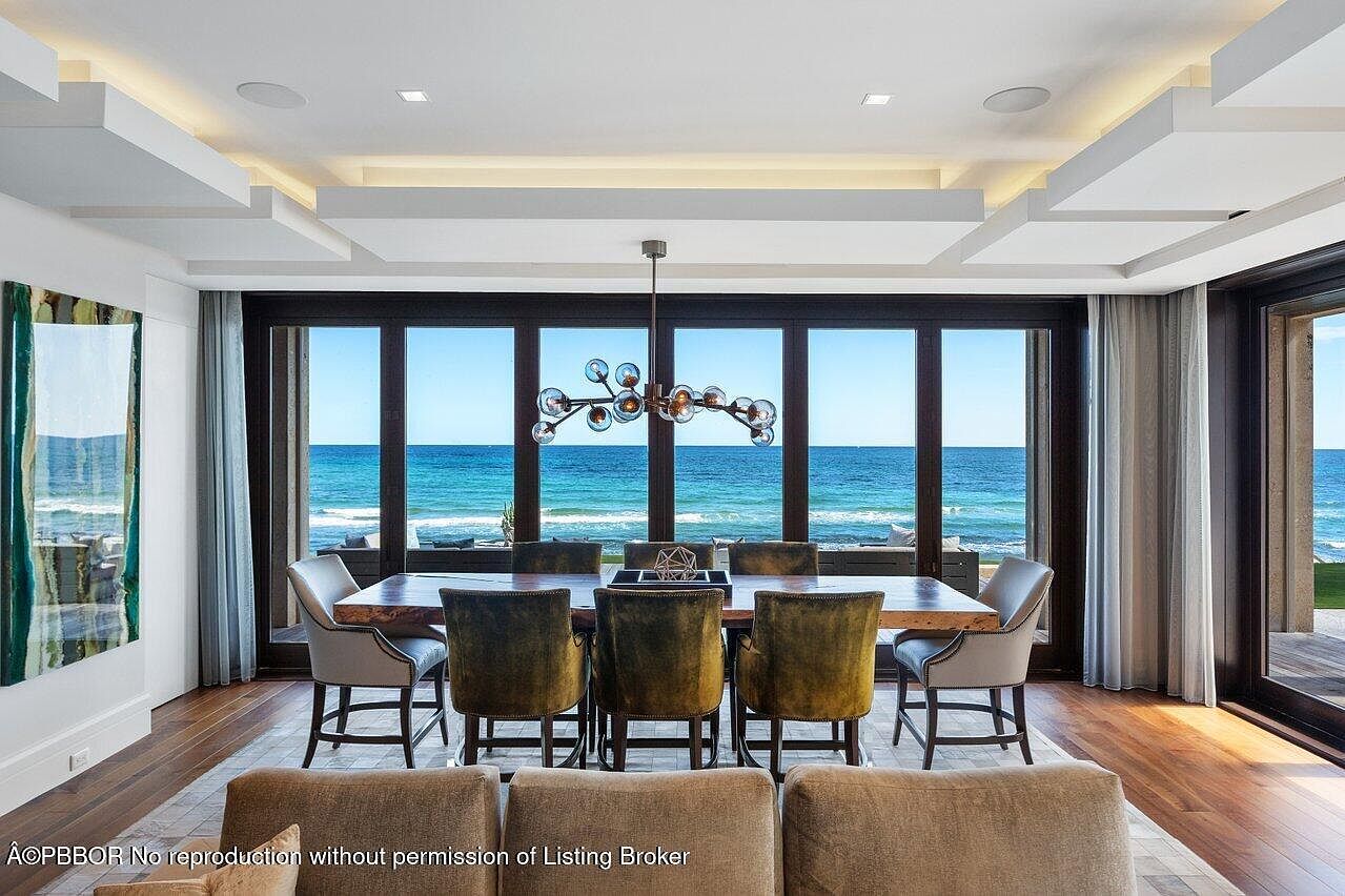 This is an interior shot of a dining room with a stunning ocean view. The room features a large wooden dining table surrounded by upholstered chairs, illuminated by a modern chandelier. The expansive windows offer an unobstructed view of the ocean, creating a luxurious and serene dining experience.