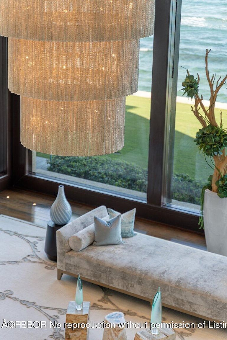 This interior shot showcases a luxurious living room with a focus on high-end design elements. A multi-tiered chandelier dominates the upper portion of the frame, while a stylish chaise lounge with decorative pillows sits below. Large windows offer a view of the ocean and manicured landscaping, enhancing the room's appeal.