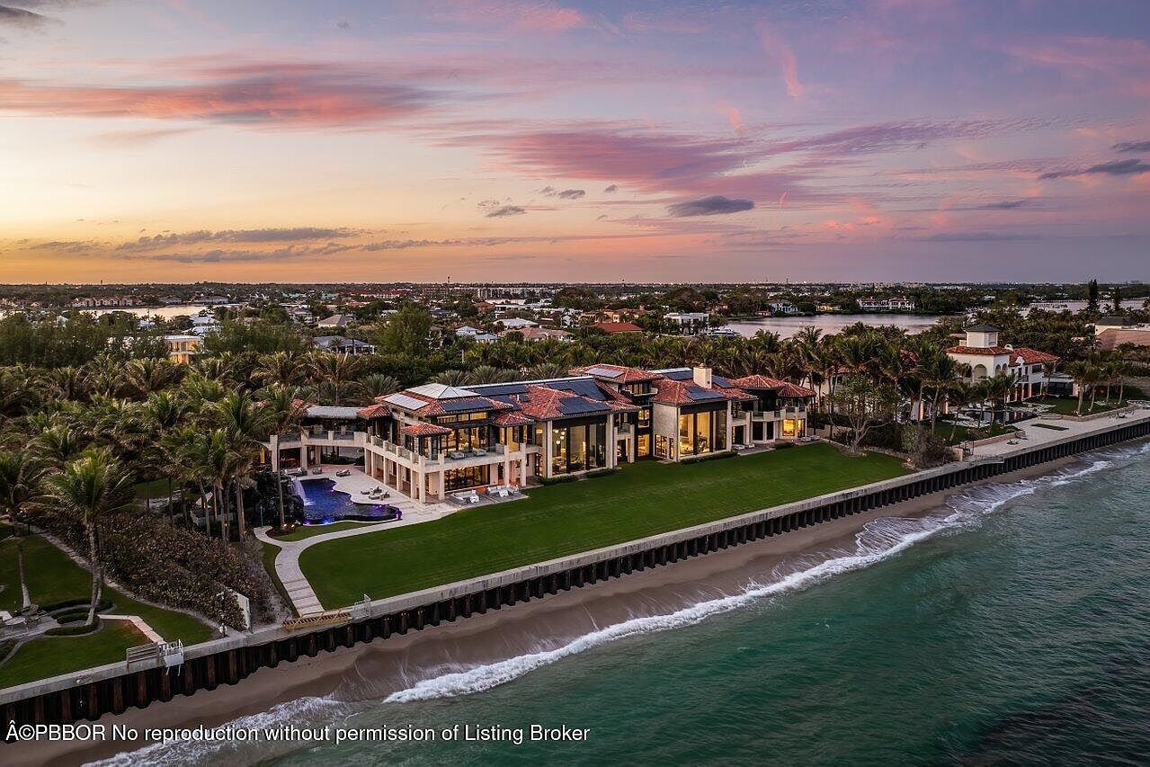 This aerial view showcases a luxurious waterfront estate with a sprawling green lawn extending to the beach. The residence features a red tile roof, multiple stories, and large windows offering stunning views. A private pool and lush landscaping add to the property's appeal, creating an impression of opulence and exclusivity.