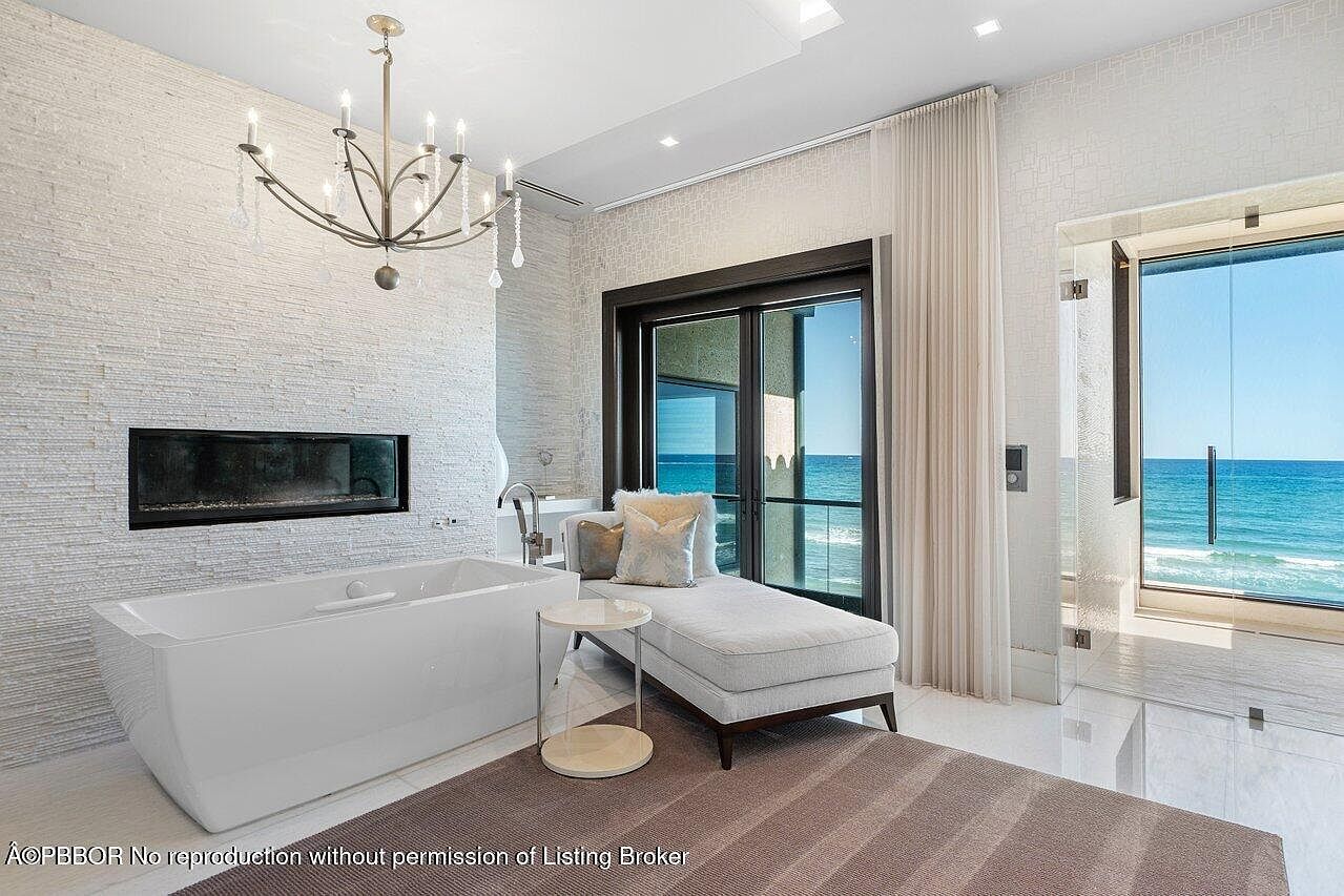 This is a luxurious primary bathroom featuring a modern freestanding bathtub positioned in front of a textured white stone wall with a built-in fireplace. A chaise lounge sits near a large sliding glass door offering ocean views, complemented by a chandelier overhead. The room exudes a spa-like ambiance with its clean lines, neutral color palette, and natural light.