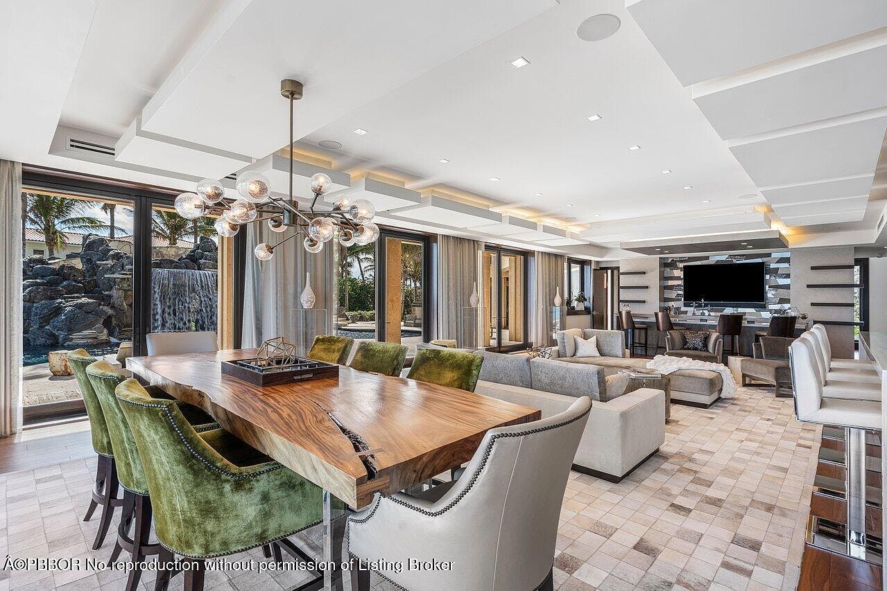 This is an interior shot of a luxurious living and dining area. The space features a large wooden dining table with green velvet chairs and a modern chandelier, seamlessly flowing into a living area with plush sofas and a bar area in the background. Large glass doors offer a view of an outdoor waterfall feature, enhancing the upscale and serene ambiance.