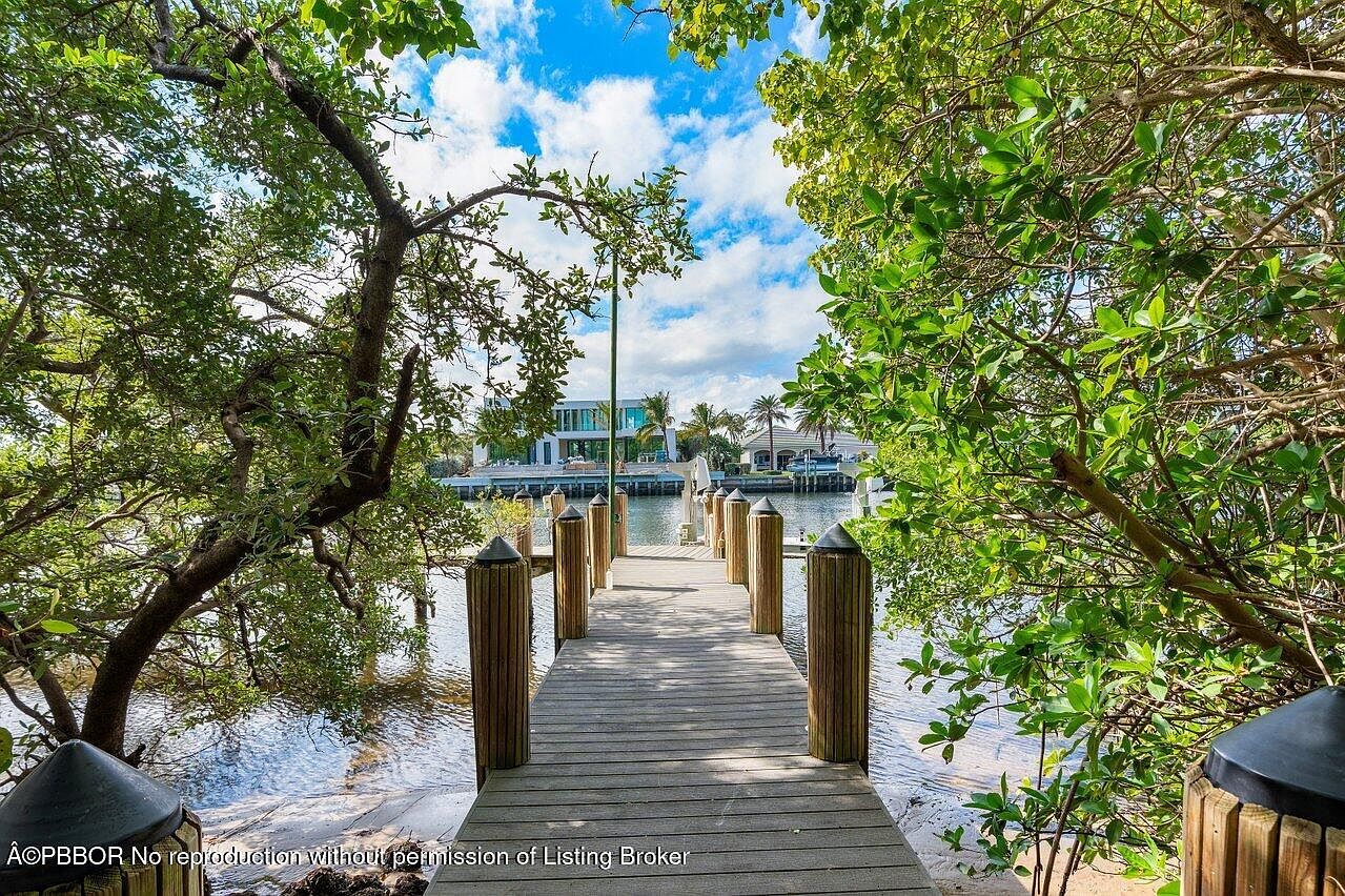 This image showcases a wooden dock extending into the water, framed by lush greenery on either side. The dock leads towards a modern waterfront property in the distance, under a partly cloudy sky. The scene evokes a sense of tranquility and private waterfront access, highlighting a desirable feature for potential buyers.
