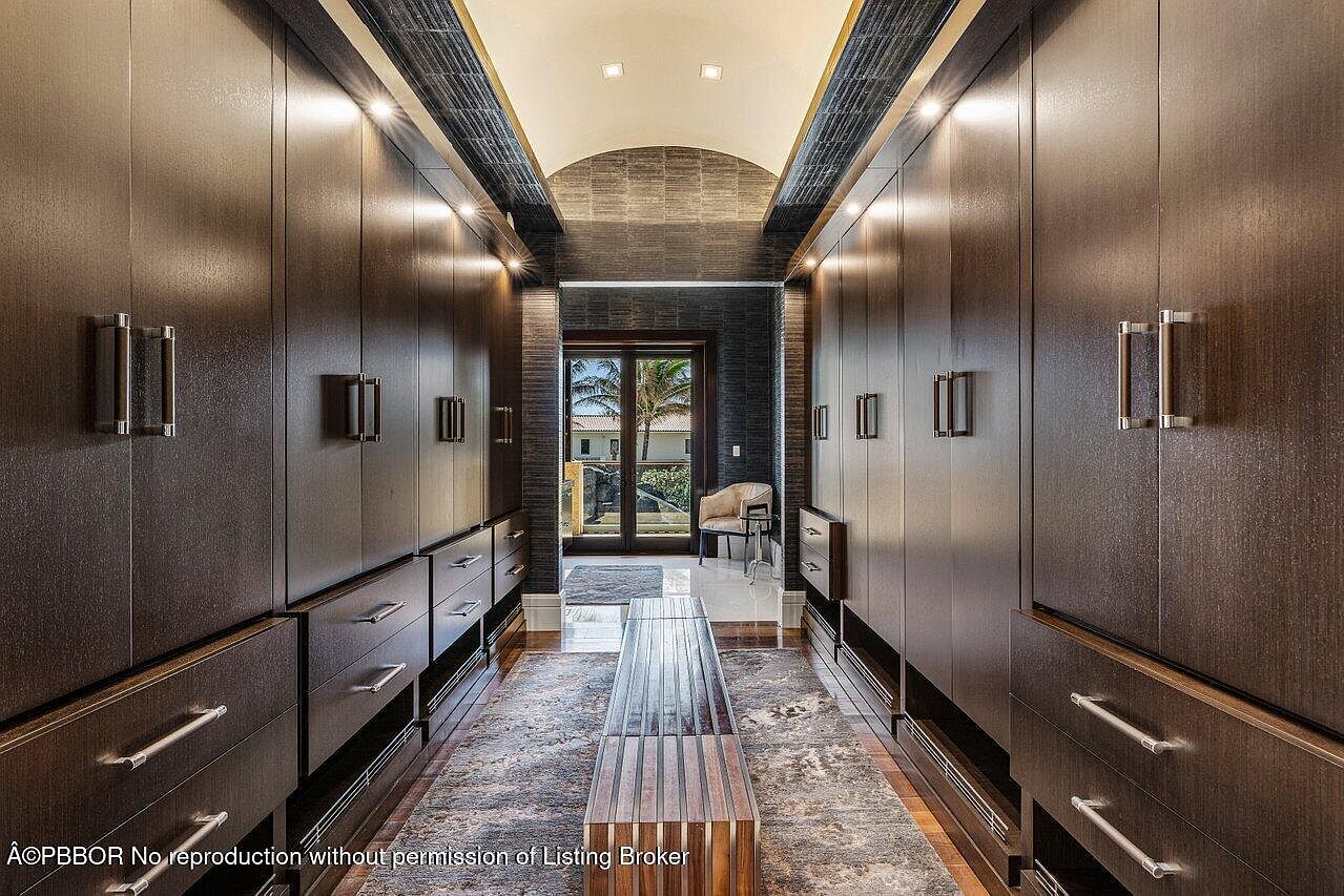 This is a well-organized walk-in closet featuring dark wood cabinetry with modern hardware. The closet has ample storage space with drawers, shelves, and hanging rods. A central bench provides a seating area, and the space is illuminated by recessed lighting, creating a luxurious and functional dressing area.