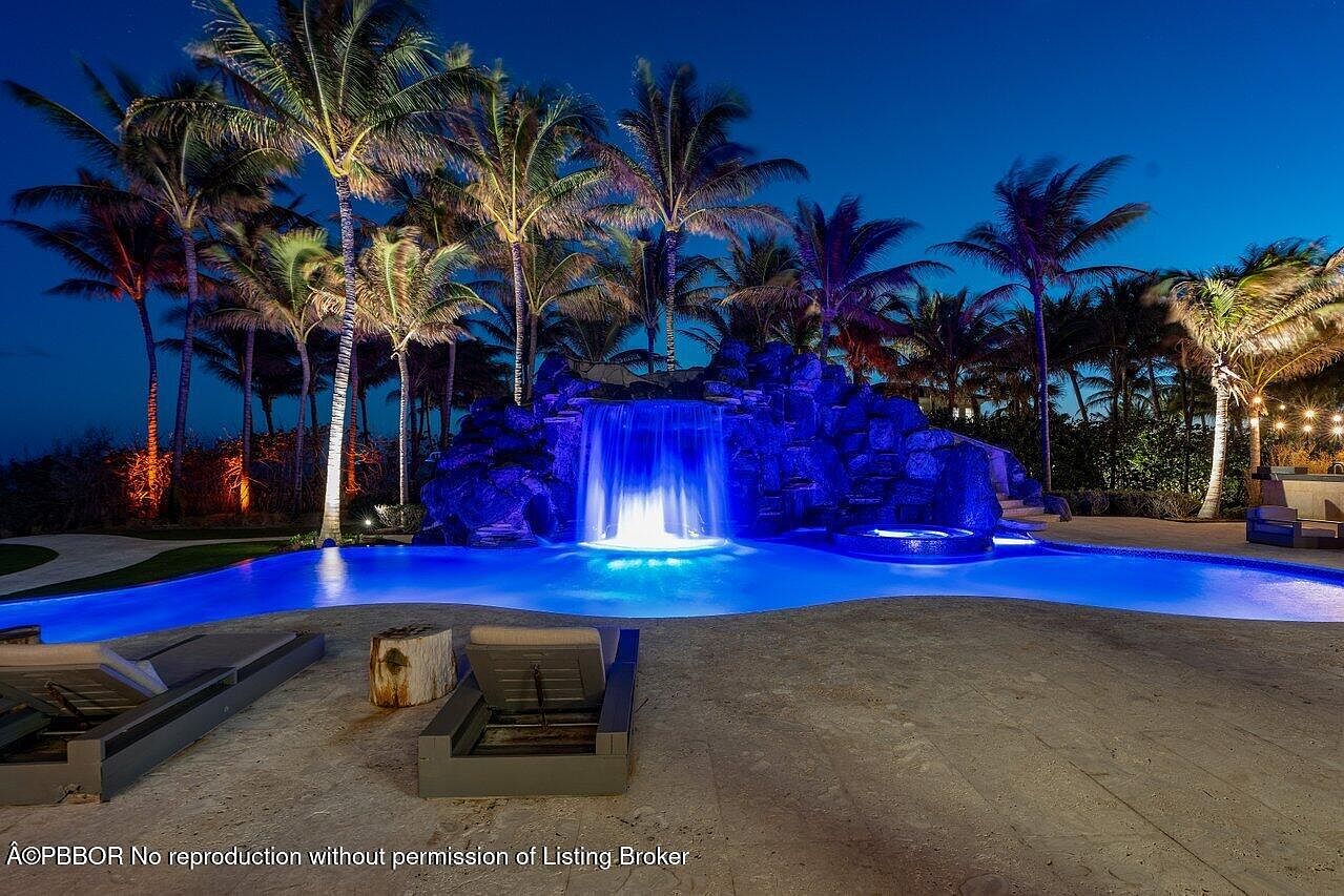 This image showcases a luxurious outdoor pool and spa area at night. The pool features a dramatic waterfall feature built into a rock formation, illuminated with blue lighting, creating a serene and inviting atmosphere. Palm trees surround the pool area, adding to the tropical ambiance, and lounge chairs are placed on the patio for relaxation.