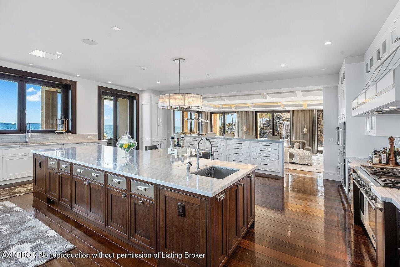 This is a spacious, high-end kitchen featuring a large island with a marble countertop and dark wood cabinetry. The kitchen boasts stainless steel appliances, a modern chandelier, and an open layout that connects to a living area with ocean views. The hardwood floors add warmth and elegance to the space.