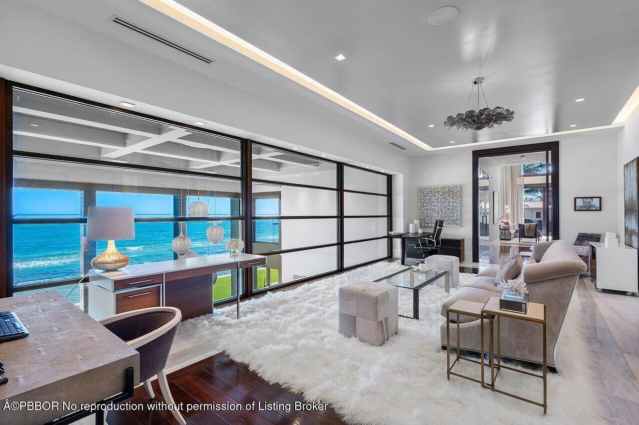 This is an interior shot of a luxurious office space with a stunning ocean view. The room features a large window overlooking the ocean, a modern desk setup, and a comfortable seating area with a plush rug. The design is contemporary with clean lines and a neutral color palette, creating a sophisticated and inviting workspace.