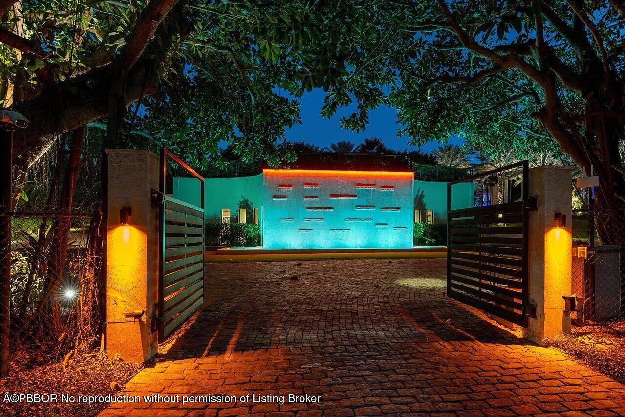 The image showcases an illuminated entryway at night, featuring an open gate leading to a property with a modern water feature wall. The gate is flanked by pillars with warm lighting, and the brick driveway adds texture to the scene. The water feature is lit with blue and orange lights, creating a striking focal point.