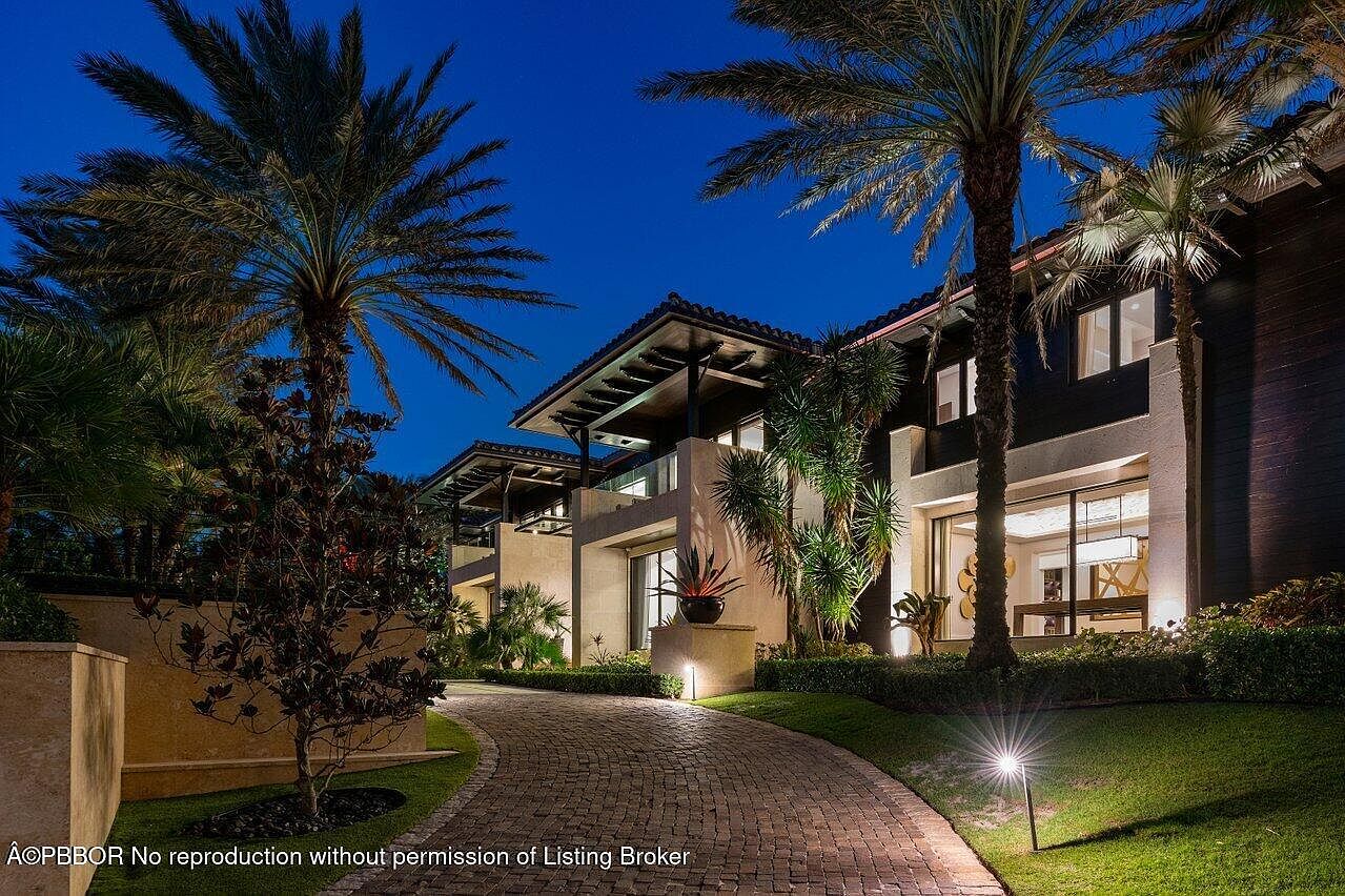 This is a front exterior view of a luxurious multi-story home at dusk. The property features lush landscaping with palm trees, manicured lawns, and a brick driveway leading to the entrance. Architectural details include a dark exterior with light-colored accents, balconies, and well-lit pathways, creating an inviting and upscale ambiance.