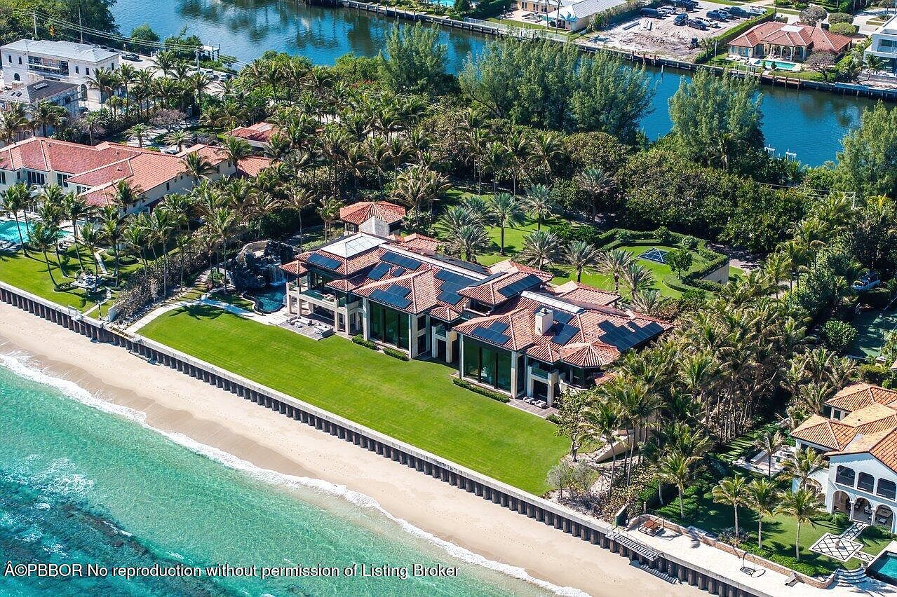 This aerial view showcases a luxurious beachfront estate with a sprawling green lawn extending to the sandy beach. The residence features a red tile roof, solar panels, and large windows, complemented by lush tropical landscaping and a private pool area. The property is situated along a picturesque coastline with clear turquoise waters.