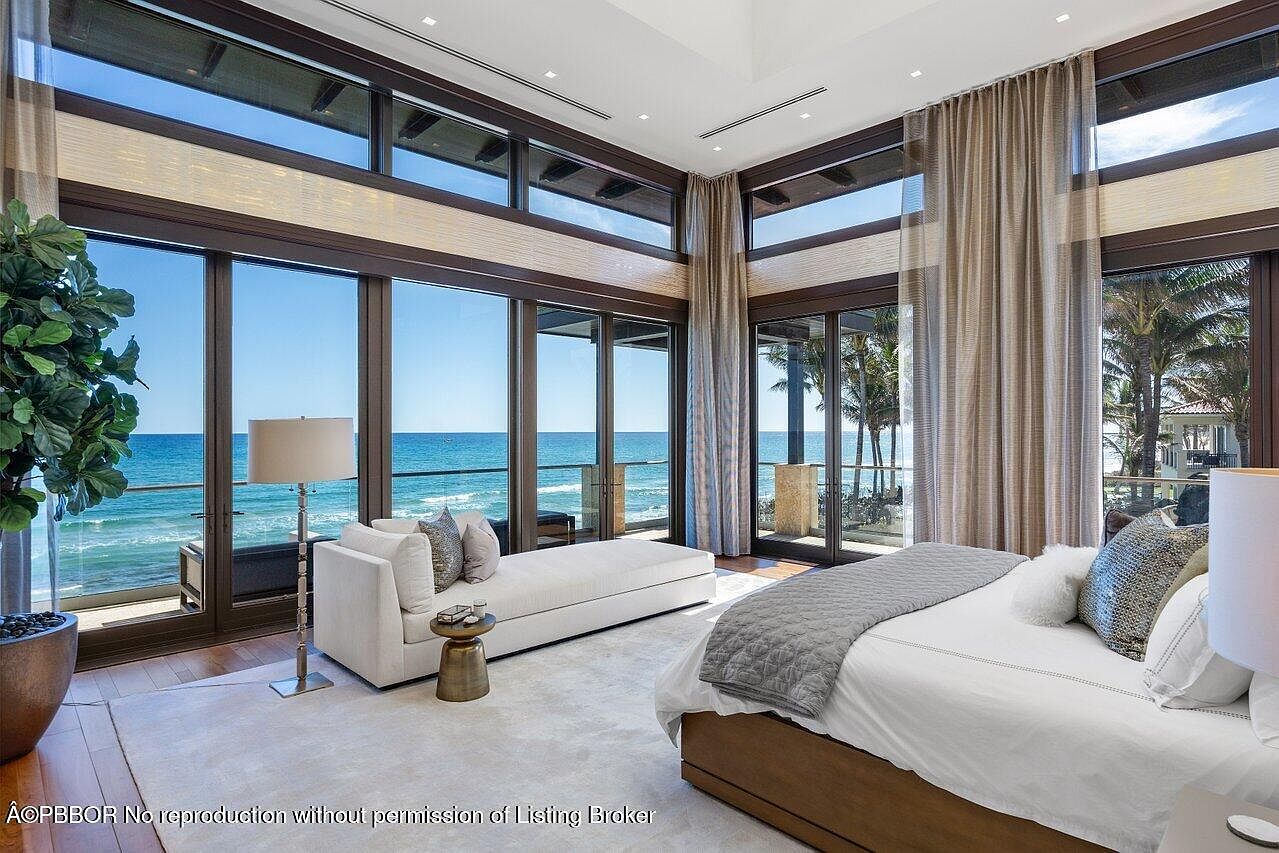This is a luxurious primary bedroom with floor-to-ceiling windows offering stunning ocean views. The room features a large bed with a gray throw, a chaise lounge, and elegant curtains. The overall style is modern and sophisticated, creating a serene and inviting atmosphere.