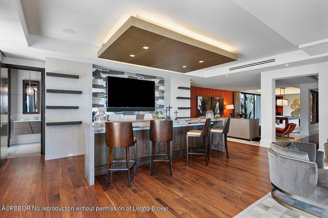 This interior shot showcases a stylish living room with a modern bar area. The bar features a dark wood counter with seating, a mirrored backsplash, and a flat-screen TV. The room is decorated with contemporary furniture and warm wood flooring, creating an inviting and luxurious atmosphere.