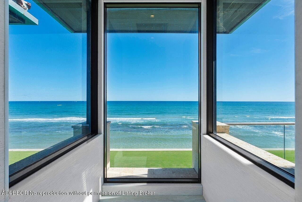 This image showcases a sunroom or conservatory with a stunning ocean view framed by large windows. The room features a minimalist design with white walls and a glimpse of green grass outside, creating a seamless transition between indoor and outdoor spaces. The expansive windows offer abundant natural light and emphasize the breathtaking coastal scenery.
