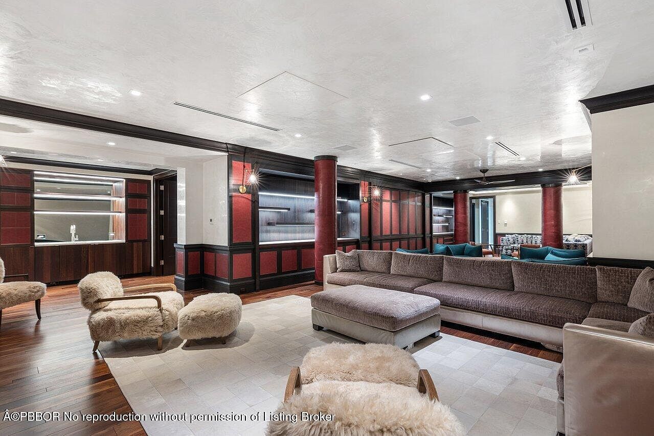 This is a spacious living room featuring a large sectional sofa, plush armchairs, and a light-colored area rug. The room has dark wood flooring and custom built-in cabinetry with red accents. The overall style is luxurious and comfortable, creating an inviting space for relaxation and entertainment.