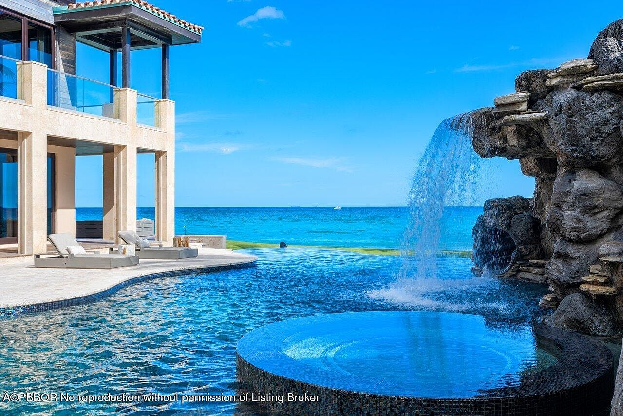 This image showcases a luxurious outdoor pool and spa area, featuring a stunning waterfall feature cascading into the pool. The pool is surrounded by a patio with lounge chairs, and a modern building with a balcony overlooks the scene. The ocean is visible in the background, creating a serene and upscale ambiance.