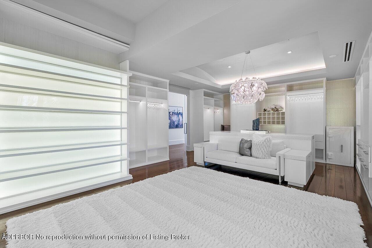 This is a spacious walk-in closet featuring custom white shelving and storage units, illuminated display areas, and a plush white rug. A modern chandelier hangs above a white sofa, creating a luxurious and organized space. The closet is well-lit and appears to be designed for high-end storage and relaxation.