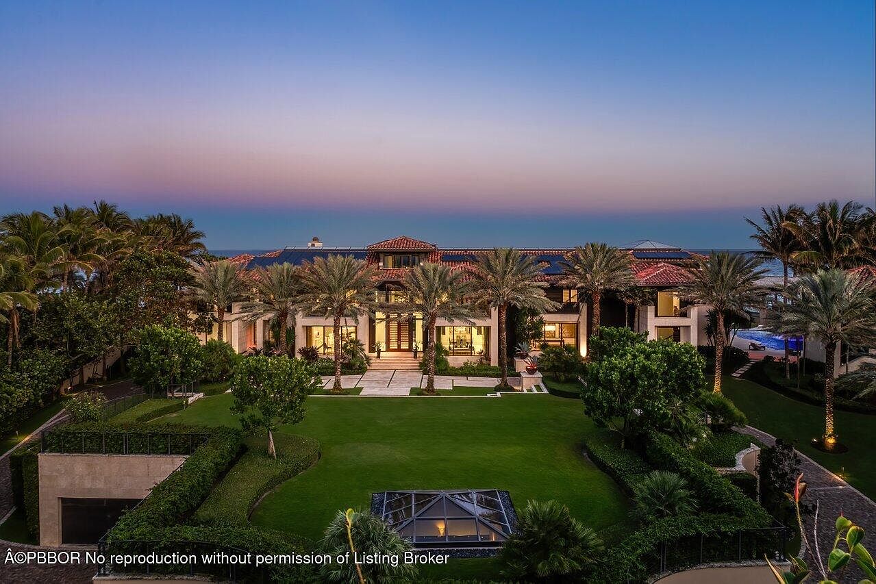 This is a stunning front view of a luxurious estate, showcasing a grand facade with multiple stories and a red tile roof. The property is beautifully landscaped with lush green lawns, mature palm trees, and manicured hedges, creating a sense of privacy and elegance. The architectural style is reminiscent of Mediterranean design, and the overall impression is one of opulence and sophistication.