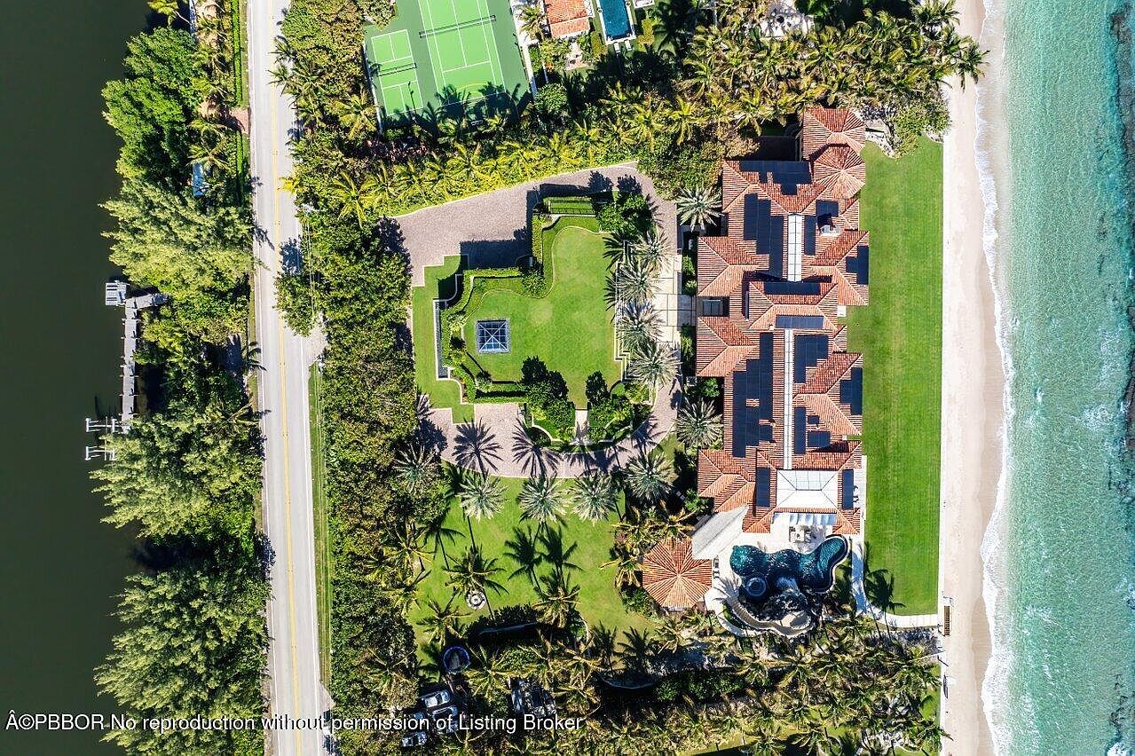 This aerial shot showcases a luxurious beachfront estate with a sprawling lawn, a large swimming pool, and direct access to a pristine beach. The property features a tennis court, meticulously landscaped gardens, and a grand residence with a terracotta tile roof and solar panels. The image highlights the property's prime location and high-end amenities.