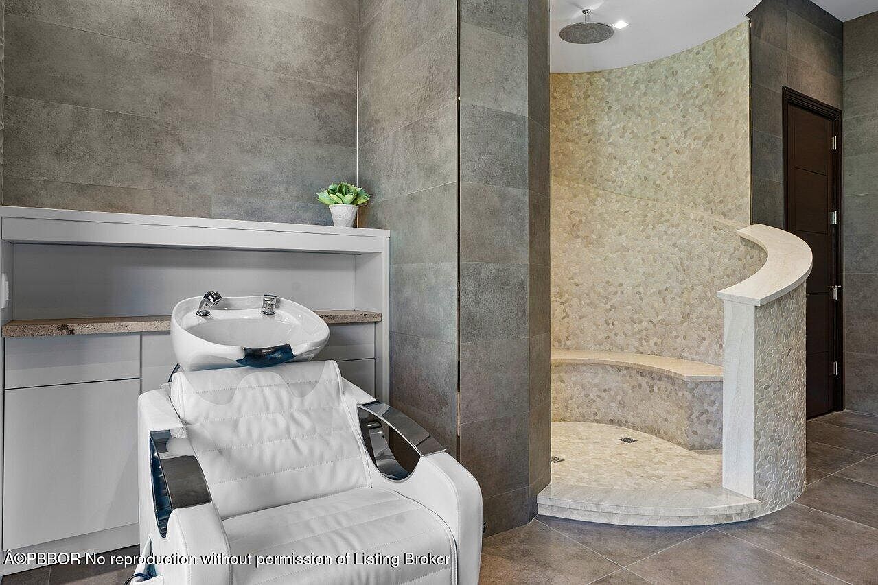 This image showcases a luxurious primary bathroom featuring a salon-style hair washing station with a comfortable white chair and a modern sink. Adjacent to the station is a unique, curved shower enclosure with pebble-style tiling and a built-in bench, creating a spa-like atmosphere. The bathroom's design incorporates sleek gray tiles and a minimalist aesthetic, emphasizing relaxation and functionality.