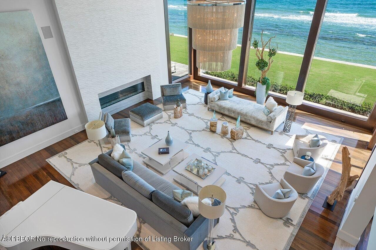 This is a high-angle shot of a luxurious living room featuring a modern fireplace, a large area rug with a floral pattern, and comfortable seating arrangements including a sofa and armchairs. Floor-to-ceiling windows offer stunning ocean views, enhancing the room's bright and airy atmosphere. A grand chandelier hangs above, adding to the room's elegance.