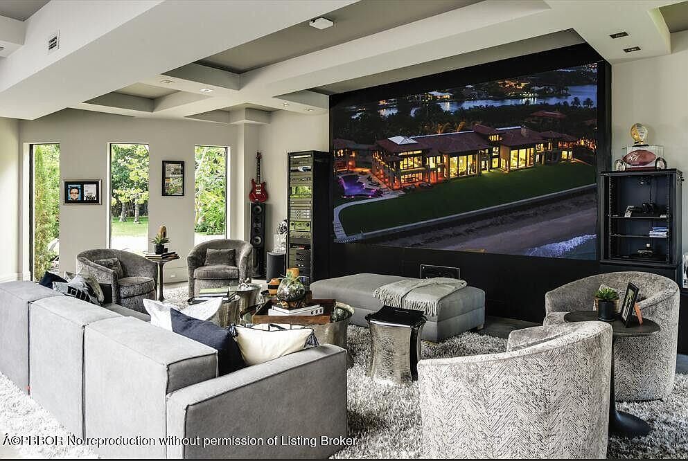 This is an interior shot of a modern living room featuring a large screen displaying a luxury home, a gray sectional sofa, and several armchairs. The room is well-lit and features a neutral color palette, creating a comfortable and inviting atmosphere. A large shag rug covers the floor, adding texture and warmth to the space.