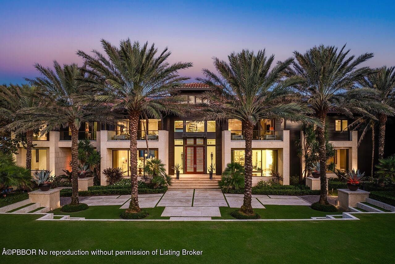 This is a front exterior view of a luxurious two-story home. The house features a symmetrical design with a central entryway, flanked by palm trees and manicured landscaping. The facade is a mix of light stone and dark wood, with large windows and balconies on the second floor, creating an inviting and upscale impression.