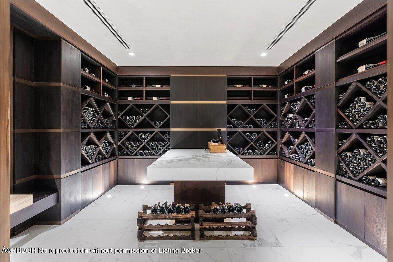This is an interior shot of a well-appointed wine cellar. The room features custom dark wood shelving with diamond-shaped bottle storage, a marble-topped center island, and recessed lighting. The overall impression is one of luxury and sophistication, perfect for showcasing a wine collection.