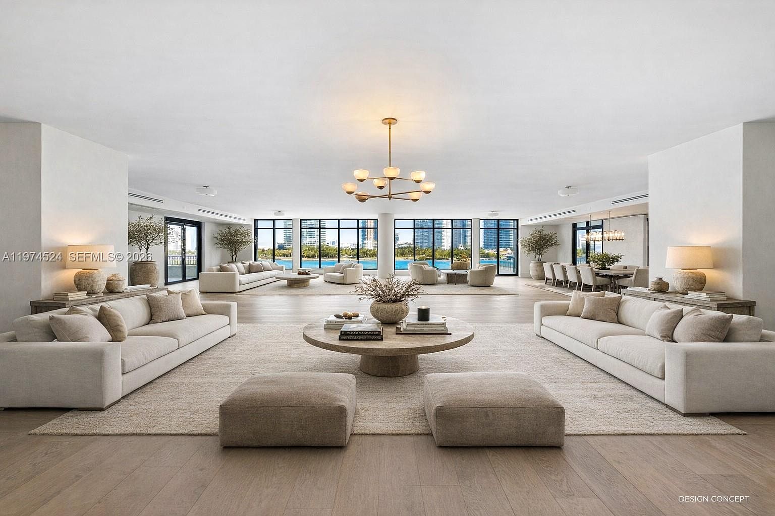 This expansive, open-concept living area features a symmetrical layout with dual beige sofas facing a central circular stone coffee table. The room is anchored by light wood flooring and large windows that provide panoramic views of the water and city skyline. A modern, multi-armed chandelier serves as a focal point, while the neutral color palette and minimalist decor create a sophisticated and airy atmosphere.