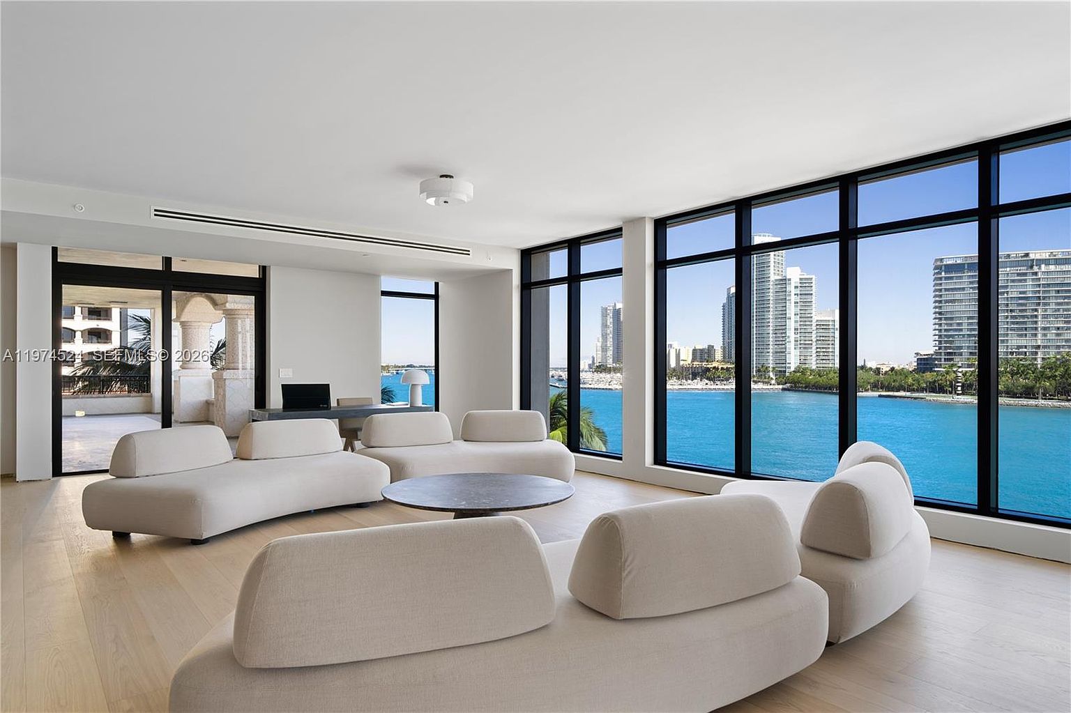 This bright and airy living room features a minimalist aesthetic with curved, light-toned sofas arranged around a circular coffee table. The space is defined by expansive, floor-to-ceiling windows that provide panoramic views of the bay and city skyline. The neutral color palette and light wood flooring create a serene, sophisticated atmosphere perfect for luxury waterfront living.