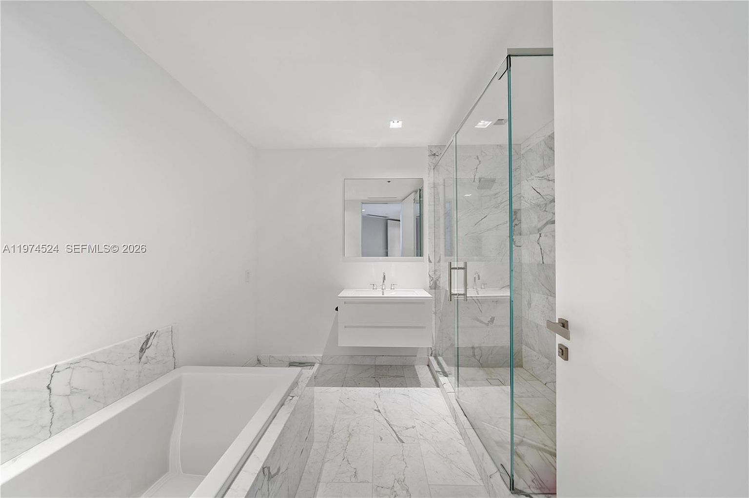 This modern primary bathroom features a clean, minimalist aesthetic with white walls and light-toned stone flooring. The space includes a deep soaking tub, a floating vanity with a rectangular sink, and a spacious walk-in shower enclosed by glass panels. The neutral color palette and bright lighting create an airy, spa-like atmosphere.