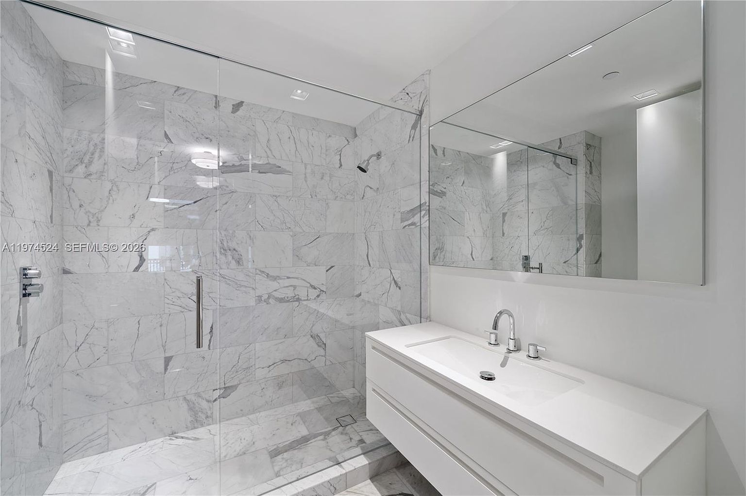 This modern bathroom features a spacious walk-in shower enclosed by a glass panel, showcasing light-toned stone tile walls and flooring. A sleek, wall-mounted white vanity with a single sink and polished chrome fixtures sits beneath a large, frameless mirror. The minimalist design creates a bright, clean, and contemporary aesthetic.