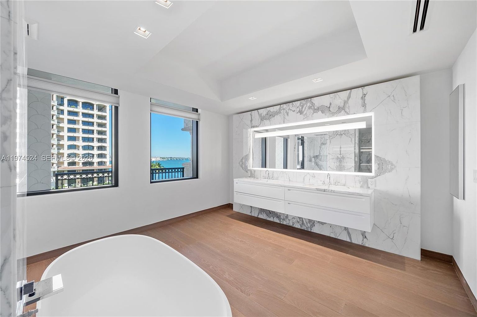 This bright and modern primary bathroom features a large, white freestanding soaking tub in the foreground and a floating double vanity with a wide, illuminated mirror. The vanity is set against a striking, large-format stone wall that adds a luxurious texture to the space. Large windows provide ample natural light and offer a clear view of the neighboring building and the water beyond.