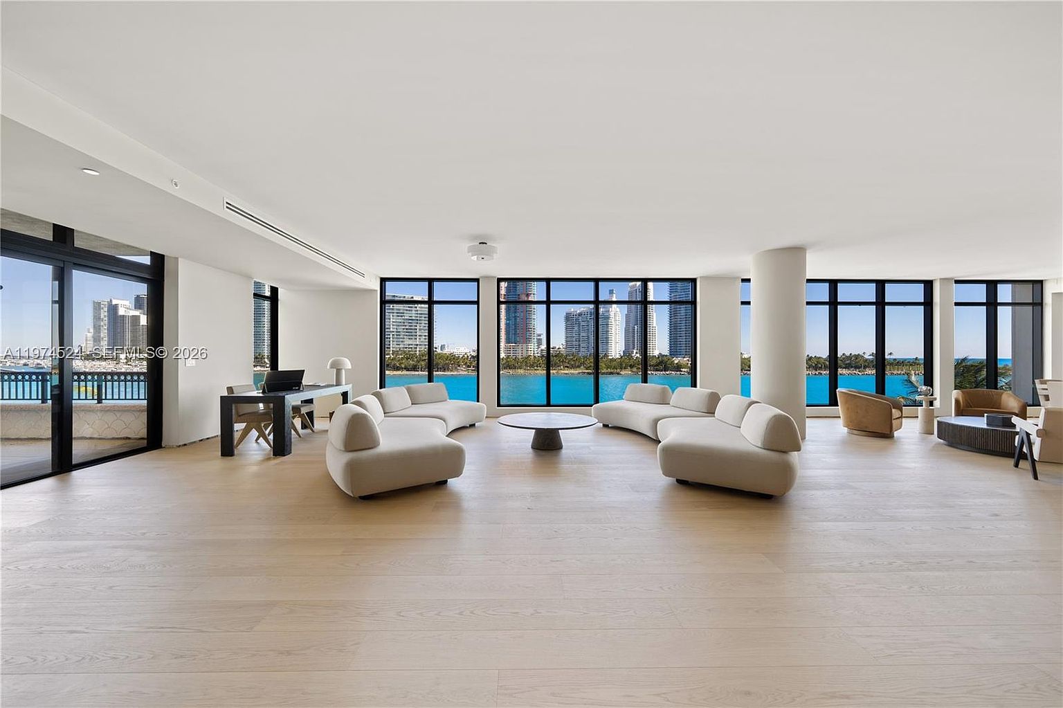 This expansive, open-concept living room features light wood flooring and a neutral color palette, centered around two large, curved sofas facing a circular coffee table. The space is defined by massive, wall-to-wall windows that provide panoramic views of the bay and city skyline, flooding the room with natural light. To the left, a sleek desk area is positioned near a sliding glass door that leads to a private balcony, creating a seamless transition between indoor and outdoor living.