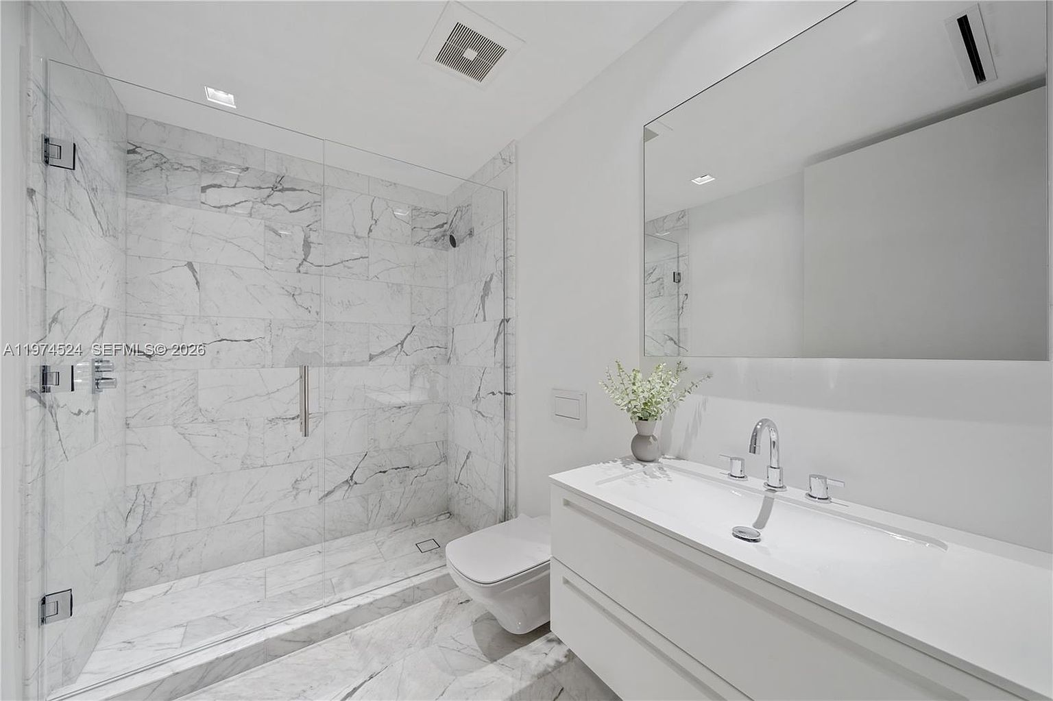 This modern, minimalist bathroom features a walk-in shower enclosed by a glass panel, showcasing light-toned stone tile with prominent veining. A sleek, wall-mounted vanity with a white countertop and integrated sink sits beneath a large, frameless mirror. The space is finished with a wall-mounted toilet and polished chrome fixtures, creating a bright and clean aesthetic.
