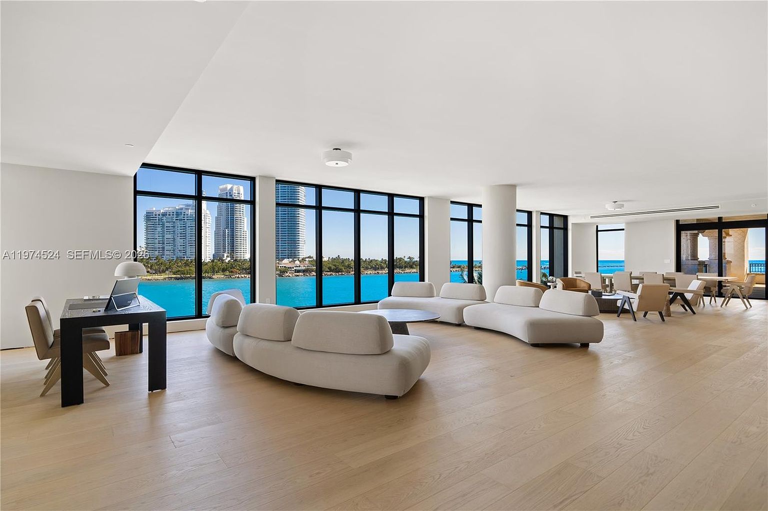 This expansive, open-concept living area features light wood flooring and a neutral color palette, anchored by two large, curved contemporary sofas. The space is defined by floor-to-ceiling windows that provide panoramic views of the water and city skyline, flooding the room with natural light. A minimalist desk setup is positioned to the left, while a dining area with modern chairs is visible in the background, creating a sophisticated and airy atmosphere.