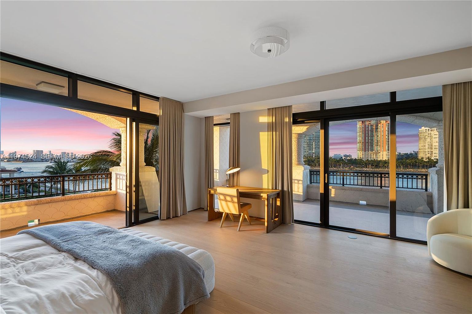 This spacious primary bedroom features light wood flooring and a neutral color palette, creating a serene and airy atmosphere. Large sliding glass doors provide access to a private balcony and offer expansive views of the water and city skyline. A dedicated workspace with a desk and chair is positioned between the glass panels, while a modern light fixture centers the room.
