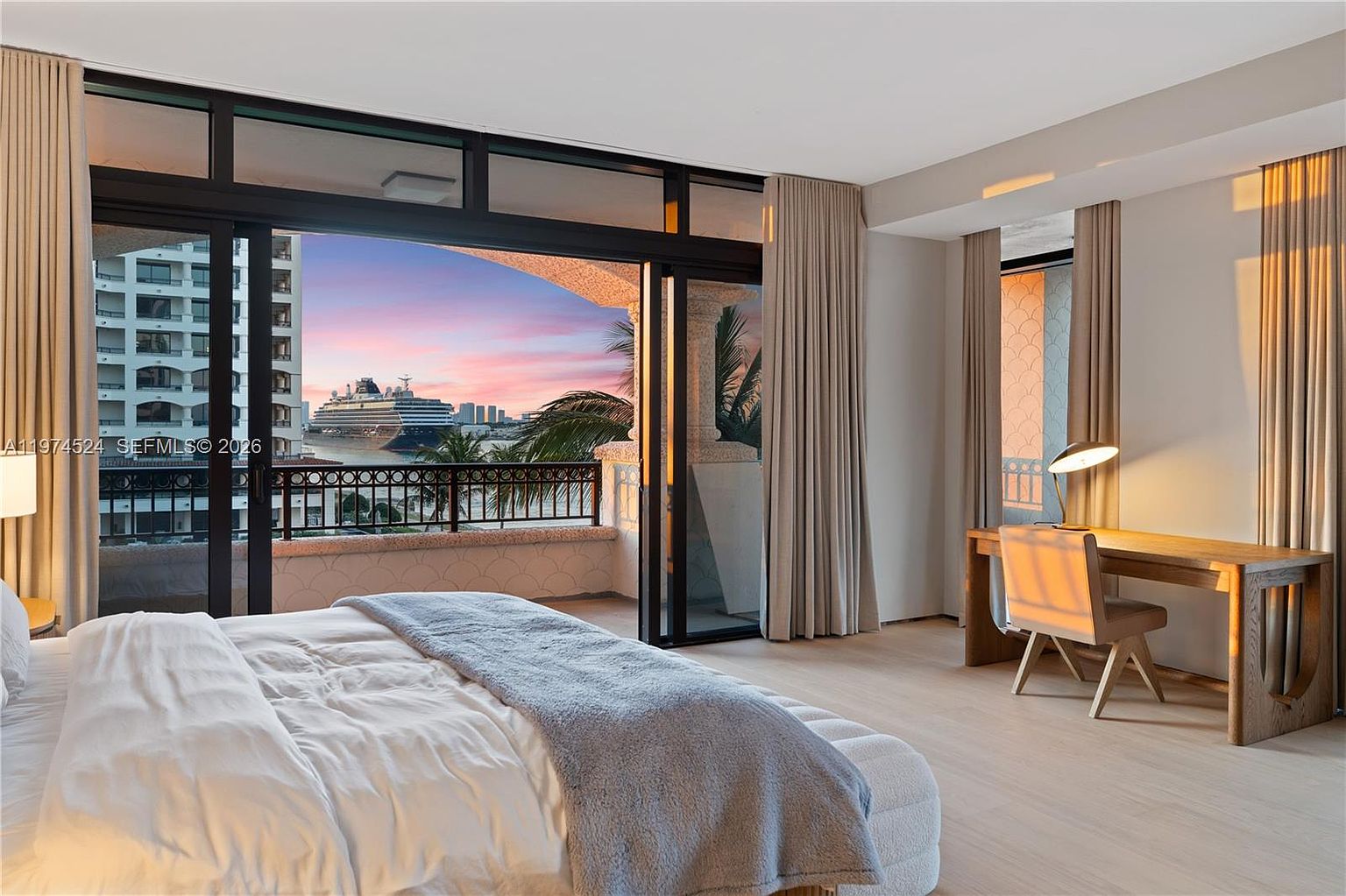 This primary bedroom features a neutral color palette and light wood flooring, creating a serene and airy atmosphere. A large sliding glass door opens to a private balcony, framing a stunning sunset view of a cruise ship in the harbor. To the right, a dedicated workspace with a desk and chair is positioned near the window, while a plush bed with a grey throw blanket anchors the room.