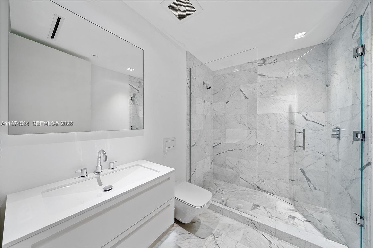 This modern bathroom features a sleek, wall-mounted vanity with a wide basin sink and a large, frameless mirror above. To the right, a spacious walk-in shower is enclosed by a glass panel and finished with light-toned, veined stone tiles. The room is illuminated by recessed ceiling lighting, creating a bright, clean, and minimalist aesthetic.