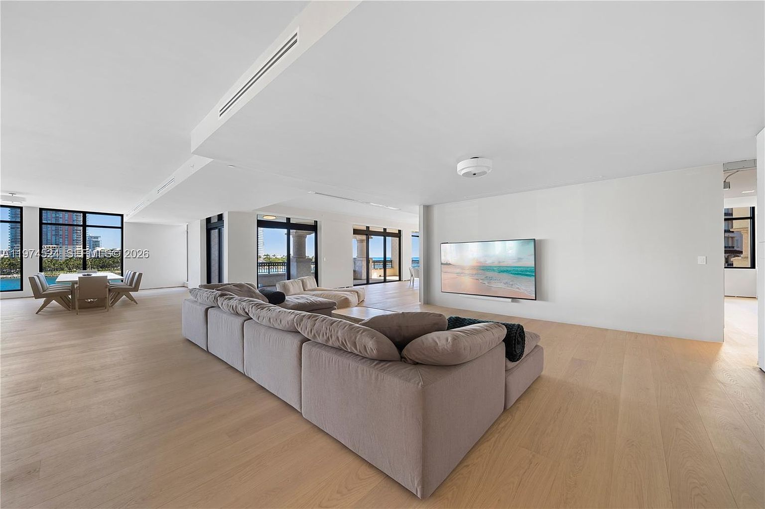 This expansive, open-concept living area features light wood flooring and a neutral color palette, creating a bright and airy atmosphere. A large, plush sectional sofa serves as the centerpiece, facing a wall-mounted television, while a dining area is visible in the background near large windows. The space is defined by its minimalist aesthetic and seamless flow, with multiple glass doors providing access to the outdoors.