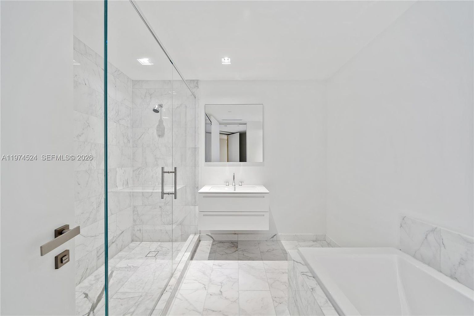This bright, contemporary bathroom features a minimalist aesthetic with white walls and light-toned stone tile flooring. The space includes a glass-enclosed walk-in shower with a rainfall showerhead, a floating vanity with a single sink, and a deep soaking tub positioned to the right. The perspective is from the doorway, looking into the clean, well-lit room that emphasizes a spa-like atmosphere.