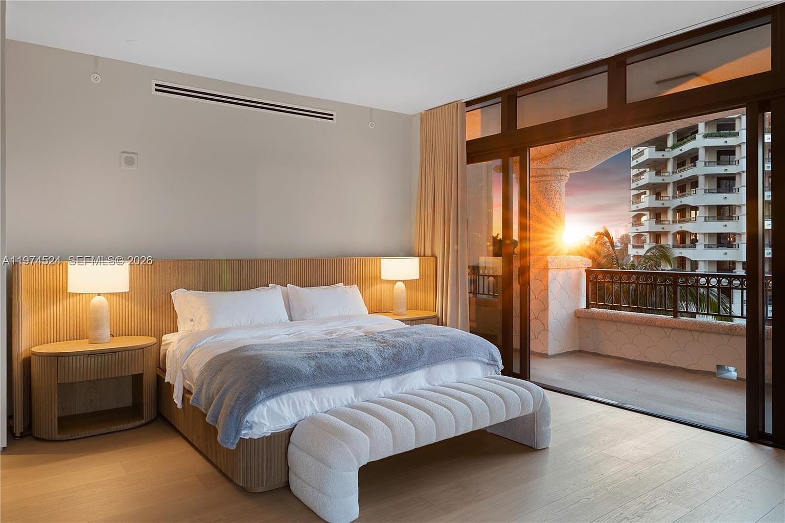 This primary bedroom features a contemporary design with a light wood-paneled headboard wall and matching bedside tables. A plush, textured bench sits at the foot of the bed, which is dressed in crisp white linens and a soft blue throw. Large sliding glass doors open onto a private balcony, allowing warm sunset light to flood the room and providing a scenic view of the neighboring building.
