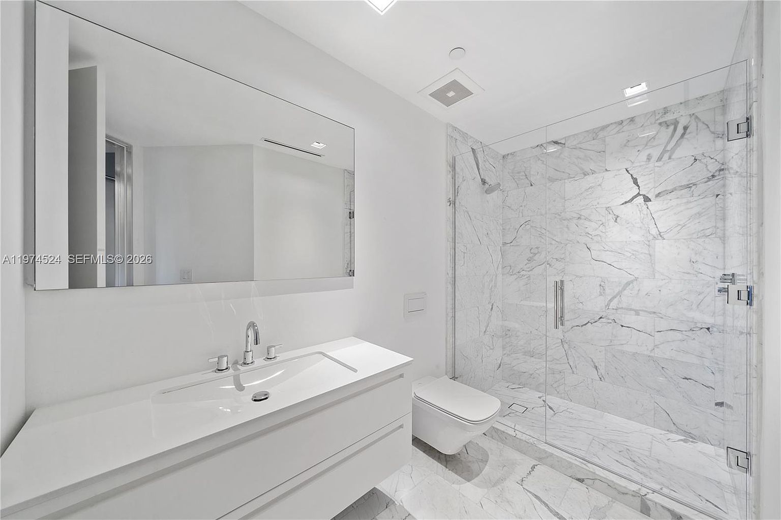 This modern, minimalist bathroom features a sleek white vanity with a rectangular sink and a large, frameless wall mirror. To the right, a glass-enclosed walk-in shower is finished with light-toned, veined stone tile that extends across the floor. The space is bright and clean, utilizing a monochromatic palette to create a contemporary and airy atmosphere.