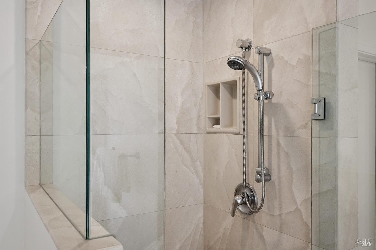 This close-up shot captures a modern shower stall featuring large, light-toned stone tiles with subtle veining. The space includes a chrome handheld shower fixture on a sliding bar and a built-in recessed niche for storage. A glass panel enclosure provides a clean, contemporary aesthetic, highlighting the minimalist design of the bathroom.