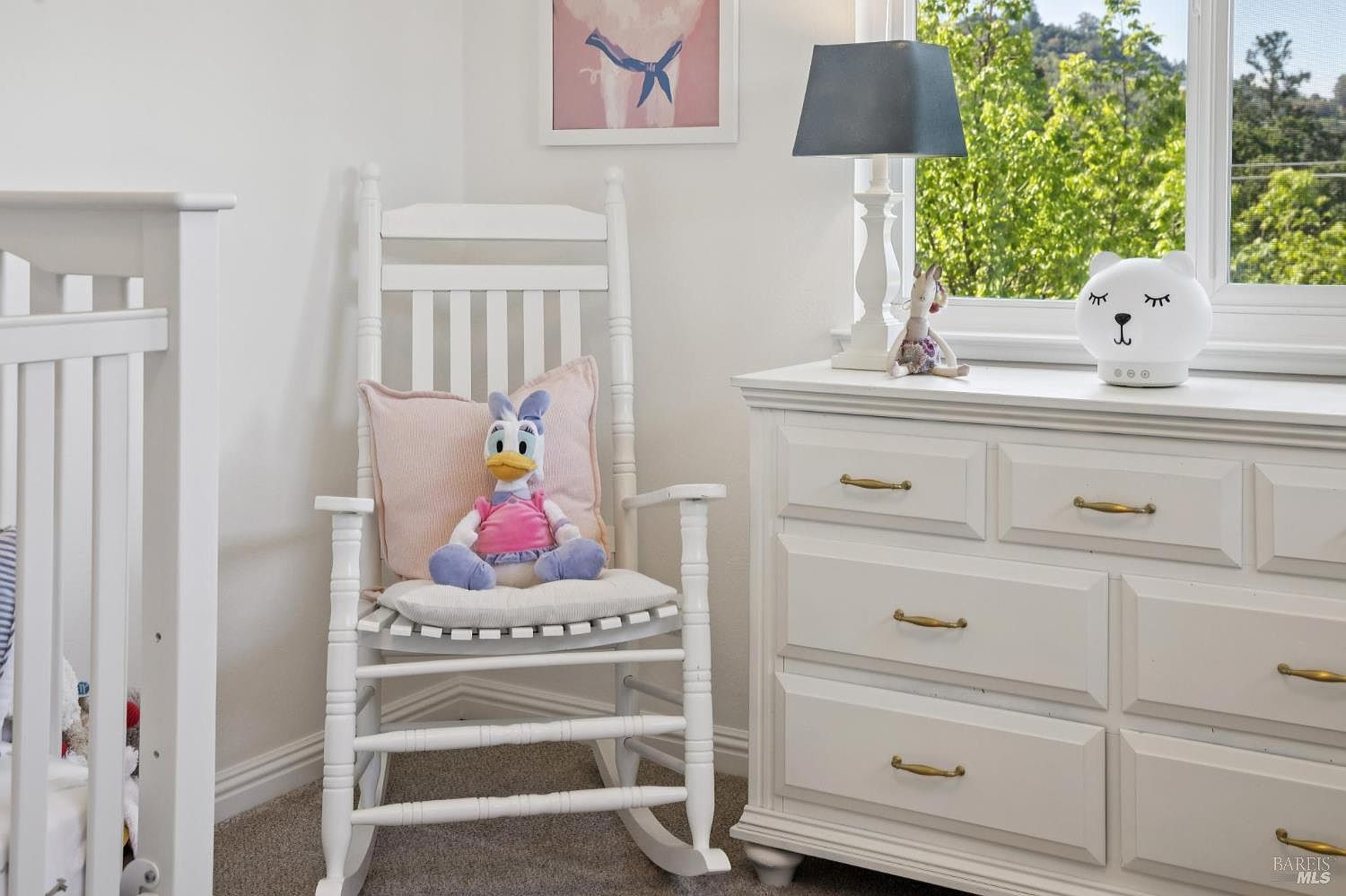 This charming nursery features a white wooden rocking chair adorned with a plush toy, positioned next to a white dresser. A lamp with a dark shade and a whimsical bear-shaped nightlight sit atop the dresser, while a framed piece of art hangs on the wall above the chair. The room is bright and inviting, with a window providing natural light and a view of the outdoors.