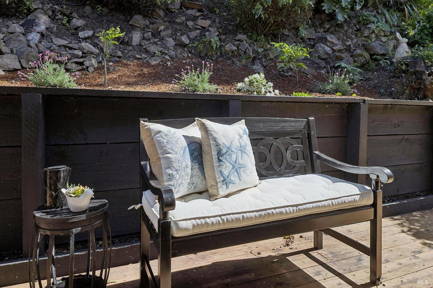 This inviting outdoor space features a dark-stained wooden bench adorned with decorative coastal-themed pillows, set upon a wooden deck. To the left, a small side table holds a potted succulent, while a tiered garden bed with lavender and small shrubs rises behind a dark wooden privacy fence. The scene offers a peaceful, well-maintained retreat with a natural, terraced landscape backdrop.