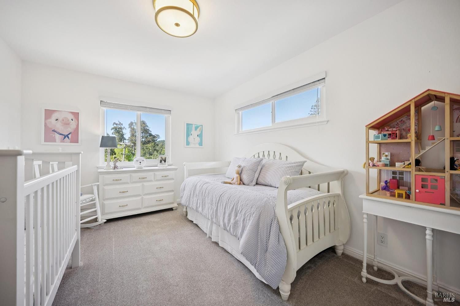 This bright and airy child's bedroom features a white daybed with striped bedding, a matching white dresser, and a crib. The room is decorated with playful wall art and a dollhouse, creating a cozy and inviting atmosphere. Large windows allow for ample natural light, highlighting the neutral carpet and clean white walls.