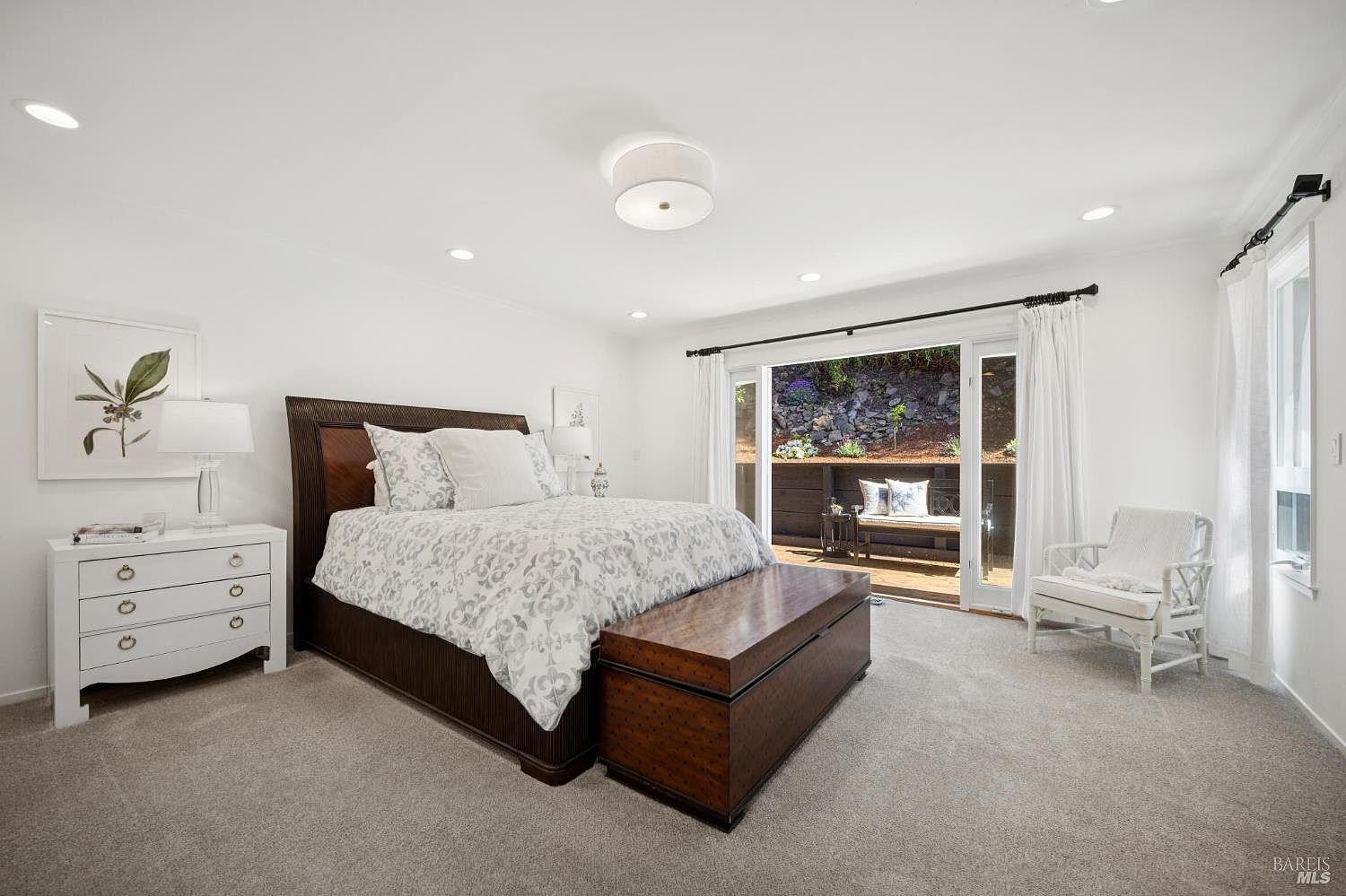 This bright and airy primary bedroom features a large dark wood bed frame with a patterned duvet and a matching wooden storage chest at the foot of the bed. A white dresser with a lamp sits to the left, while a white wicker chair is positioned near the large sliding glass doors that open onto a wooden deck. The room is decorated in a neutral palette with soft carpet and crisp white walls, creating a serene and inviting atmosphere.